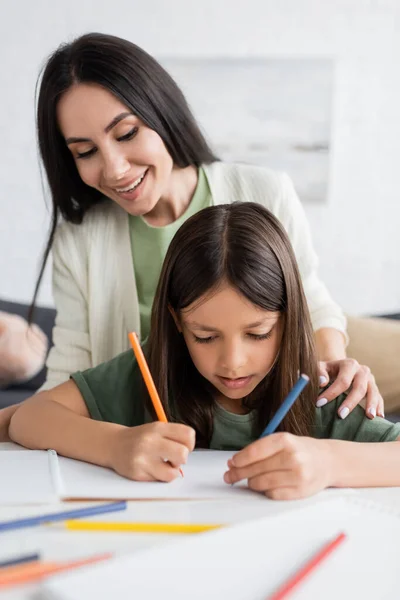 Happy babysitter looking at cheerful kid drawing on paper with colorful pencils — Stockfoto