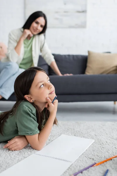 Dreamy girl lying on carpet and thinking near blurred nanny sitting on sofa — Stock Photo