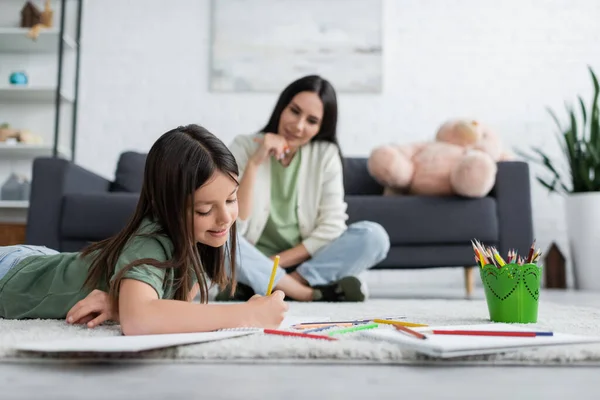 Allegra ragazza sdraiata su tappeto e disegno vicino tata offuscata seduta sul divano — Foto stock