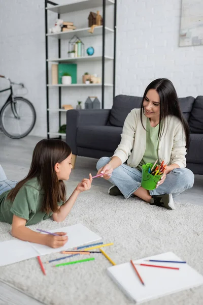 Cheerful nanny passing pencil to girl lying on carpet near blank paper — Foto stock