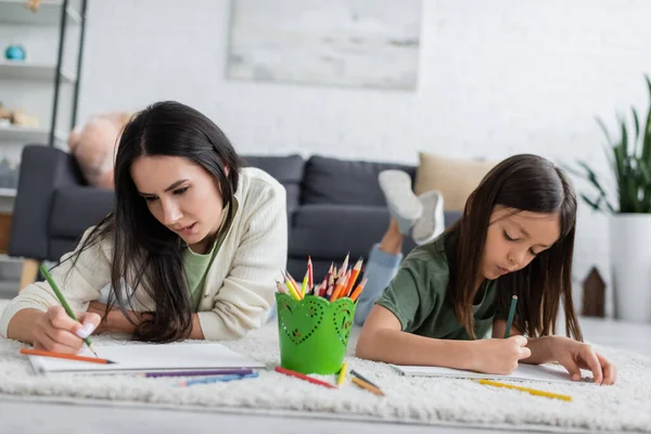 Pensive nanny biting lip near girl drawing on paper while lying on carpet in living room — Stockfoto