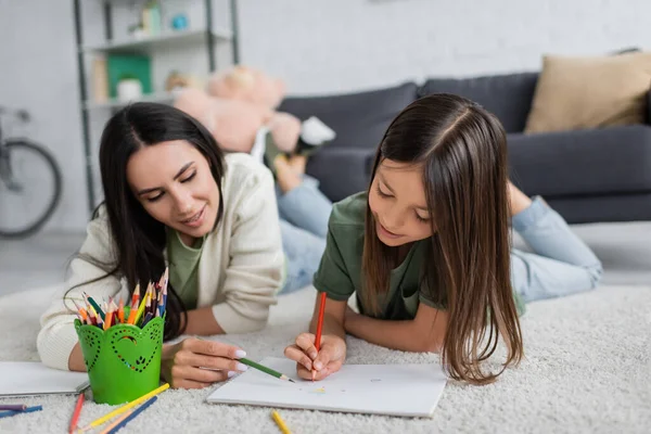 Smiling nanny and girl drawing on paper while lying on carpet in living room — Stock Photo