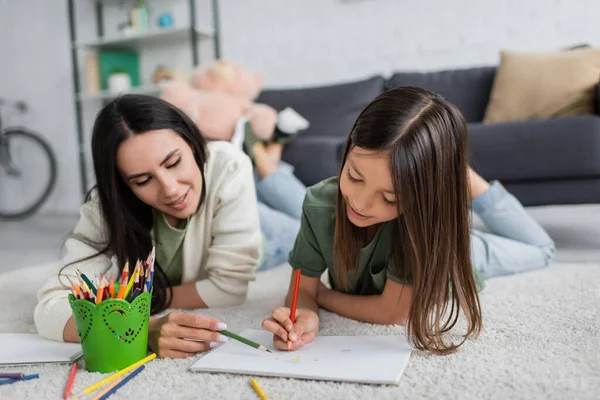 Smiling nanny and girl drawing on paper while lying on carpet in living room — Photo de stock