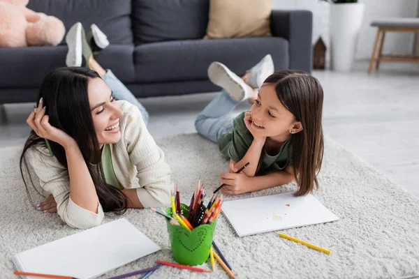 Smiling nanny and girl looking at each other while lying on carpet near blank papers in living room - foto de stock