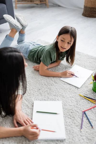 Chica acostada en la alfombra y sosteniendo lápiz cerca de papel mientras mira a la niñera borrosa - foto de stock