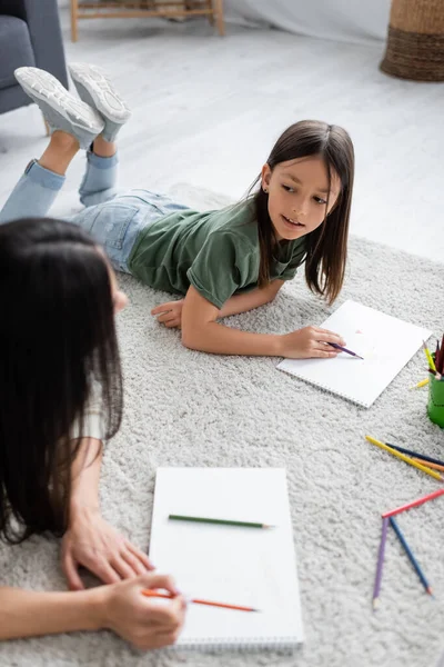 Ragazza sdraiata sul tappeto e tenendo la matita vicino alla carta mentre guardava la tata offuscata — Foto stock