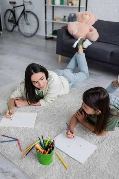 Smiling babysitter drawing and looking at paper near girl in living room - foto de stock