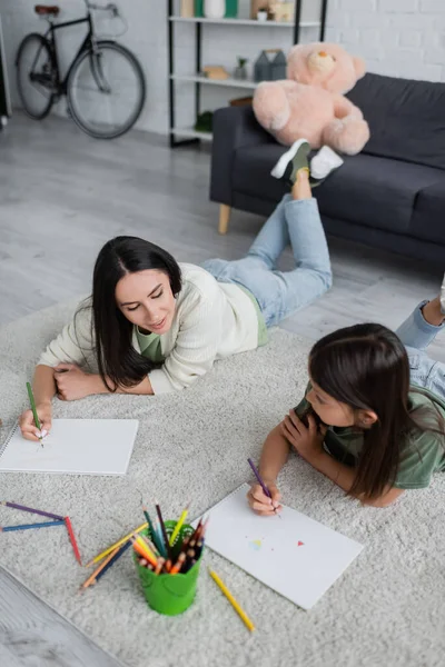 Smiling babysitter drawing and looking at paper near girl in living room — Fotografia de Stock