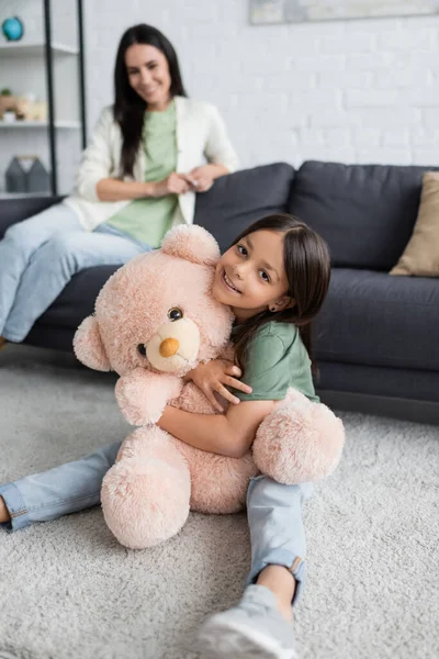 Menina feliz sentado no tapete e abraçando ursinho de pelúcia perto de babá borrada na sala de estar — Fotografia de Stock