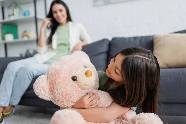 Felice ragazza guardando e abbracciando orsacchiotto vicino babysitter offuscata in soggiorno — Foto stock