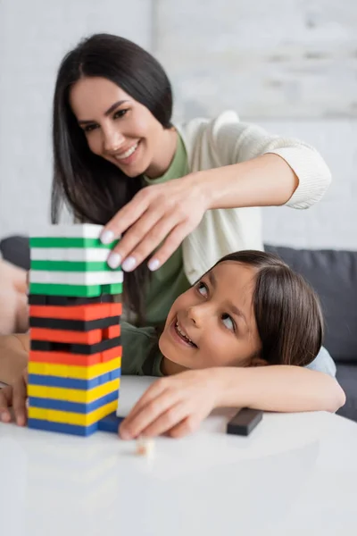 Cheerful babysitter playing wood blocks game with pleased girl in living room - foto de stock