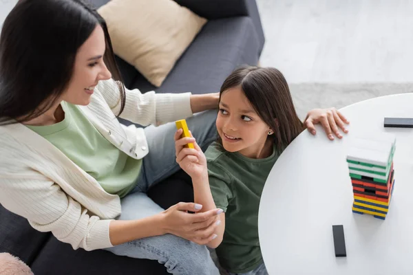 Top view of happy babysitter playing wooden tower game with positive girl in living room — Fotografia de Stock