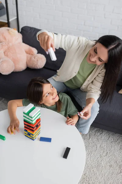 Vista ad alto angolo di babysitter positiva giocare gioco torre di legno con la ragazza felice in soggiorno — Foto stock