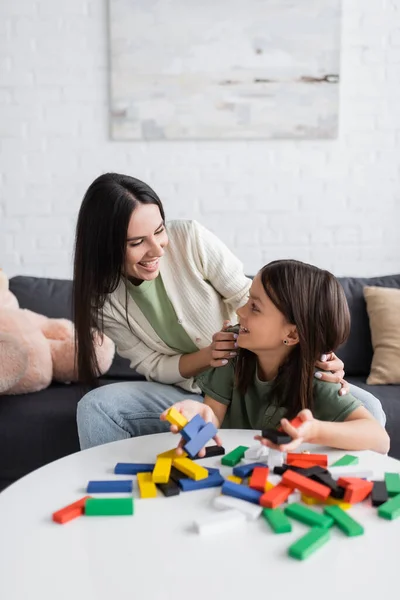 Fröhliche Babysitterin schaut Mädchen in der Nähe von bunten Holzklötzen auf dem Tisch an — Stockfoto