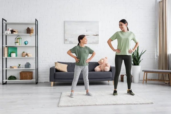 Full length of positive nanny and child standing with hands on hips while exercising at home — Stock Photo