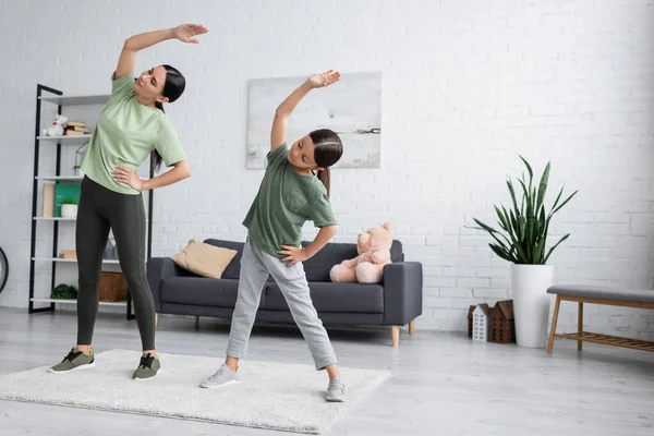 Full length of girl with babysitter doing side bend exercise with hands on hips and raised hands - foto de stock