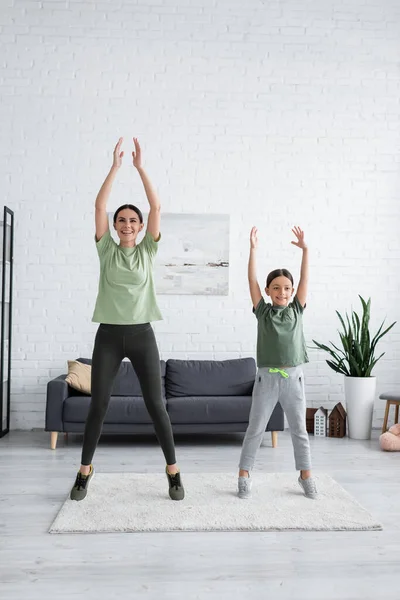 Happy girl and nanny stretching on tiptoes with raised hands in living room - foto de stock