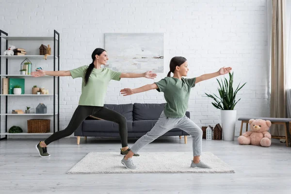 Smiling child and babysitter training in warrior pose in modern living room — Stockfoto