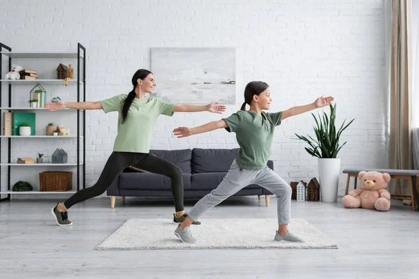 Souriant enfant et baby-sitter formation dans la pose guerrier dans le salon moderne — Photo de stock