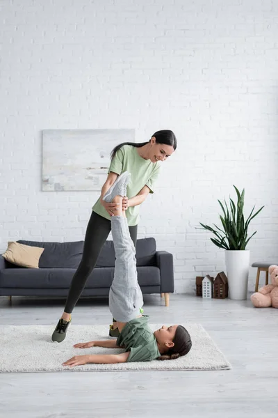 Smiling nanny helping girl exercising in supported shoulder stand pose on carpet - foto de stock