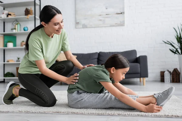 Smiling nanny assisting girl exercising in seated forward bend pose on carpet at home — Photo de stock