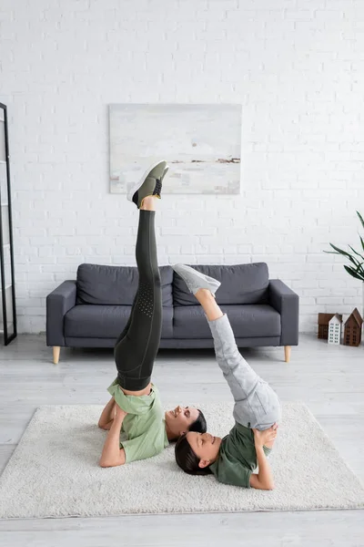 Happy girl with babysitter training in supported shoulder stand pose on carpet in living room - foto de stock