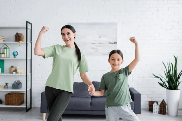 Cheerful girl and babysitter holding hands and showing muscles in living room — Stock Photo