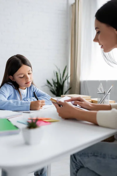 Child writing dictation near nanny reading aloud on blurred foreground — Stock Photo