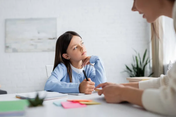 Fille rêveuse regardant loin tout en faisant des devoirs près de babysitter floue — Photo de stock