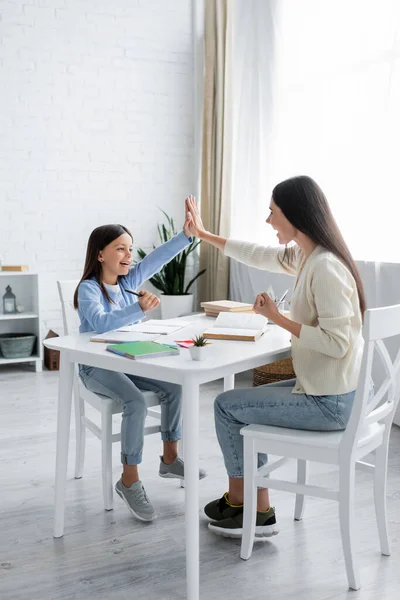 Chica emocionada y niñera dando cinco altos y mostrando el gesto de ganar mientras hacen la tarea juntos - foto de stock