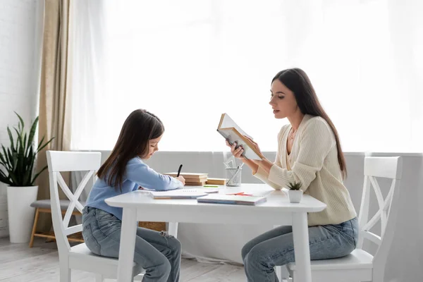 Боковой вид няни, читающей вслух рядом с девочкой, пишущей диктант дома — стоковое фото