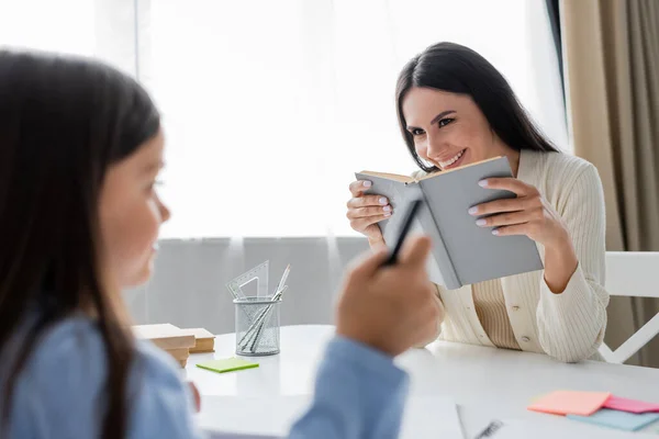 Fröhliches Kindermädchen mit schulbuchnahem Mädchen, das mit dem Finger auf verschwommenen Vordergrund zeigt — Stockfoto