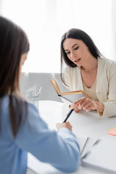 Babysitter mit Lehrbuch zeigt nahe Mädchen schreiben auf verschwommenen Vordergrund — Stockfoto