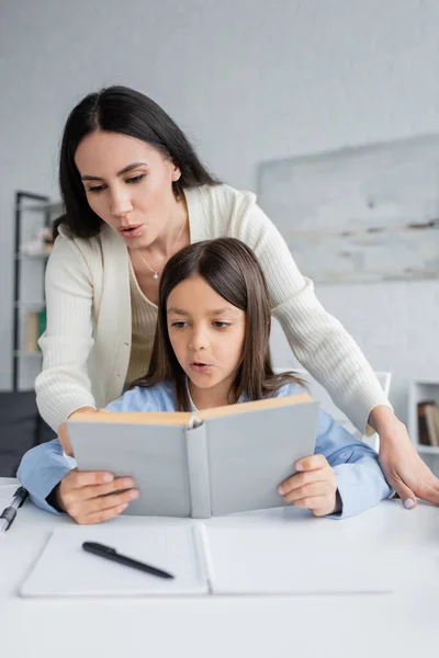 Brunette nounou aider fille lecture livre tout en faisant des devoirs — Photo de stock