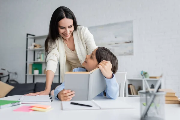 Tata sorridente vicino bambino seduto con libro di testo vicino a libri di copia sul tavolo — Foto stock