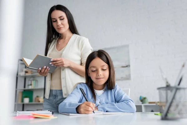 Babysitter standing with book near girl writing in copybook at home — Stock Photo