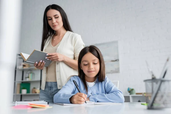 Babysitter standing with book near girl writing in copybook at home — Photo de stock