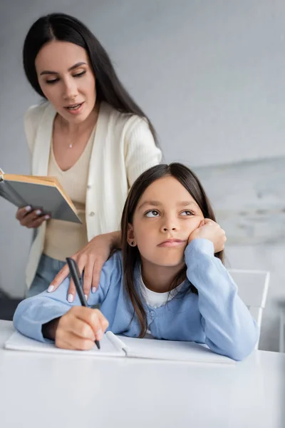 Nanny with book talking to bored girl sitting with pen near notebook — Photo de stock