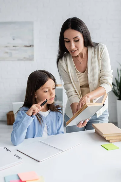 Brunette nanny showing textbook to girl sitting with pen near copybook — Foto stock