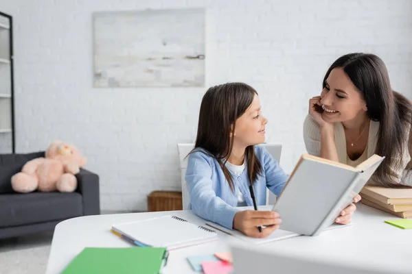 Glückliche Kindermädchen schauen Mädchen an, die mit Lehrbuch zu Hause sitzen — Stockfoto