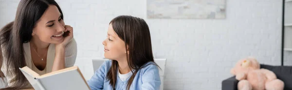 Cheerful nanny looking at girl reading book at home, banner — Stock Photo