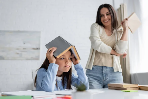 Annoiato ragazza che copre la testa con libro di testo vicino bambinaia sorridente su sfondo sfocato — Foto stock