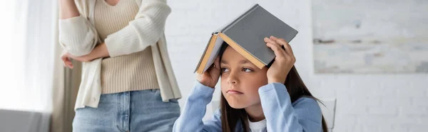 Chica aburrida y cansada cubriendo la cabeza con libro de texto cerca de niñera en el fondo, pancarta - foto de stock