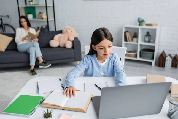 Ragazza che fa i compiti vicino al computer e bambinaia seduto con libro su sfondo sfocato — Foto stock