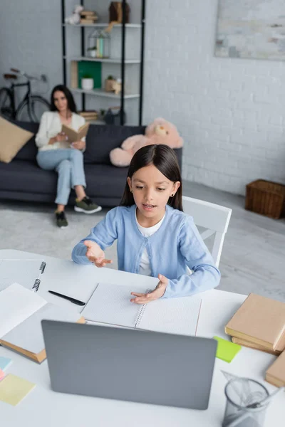 Girl talking at laptop during online lesson near babysitter on blurred background — Photo de stock