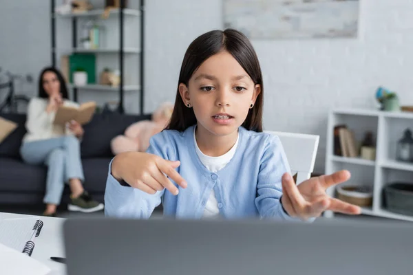 Fille pointant vers ordinateur portable pendant la leçon en ligne près de nounou sur fond flou — Photo de stock