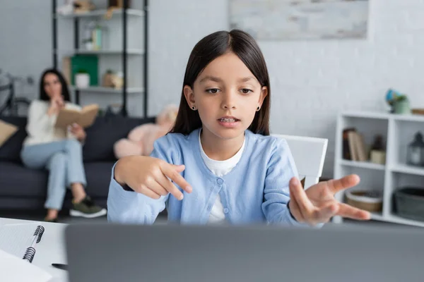 Menina apontando para laptop durante aula on-line perto de babá no fundo embaçado — Fotografia de Stock