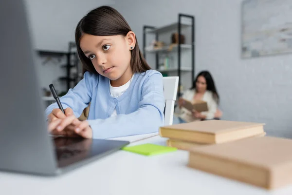Девушка пишет рядом размытый ноутбук во время выполнения домашней работы рядом с няней на заднем плане — стоковое фото