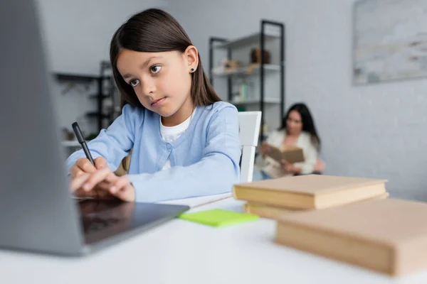 Ragazza scrittura vicino computer portatile offuscata mentre fa i compiti vicino bambinaia sullo sfondo — Foto stock