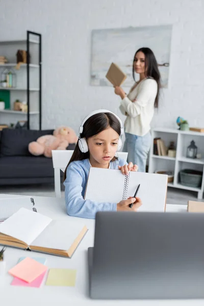 Girl in headphones pointing at blank notebook near laptop and nanny on blurred background - foto de stock